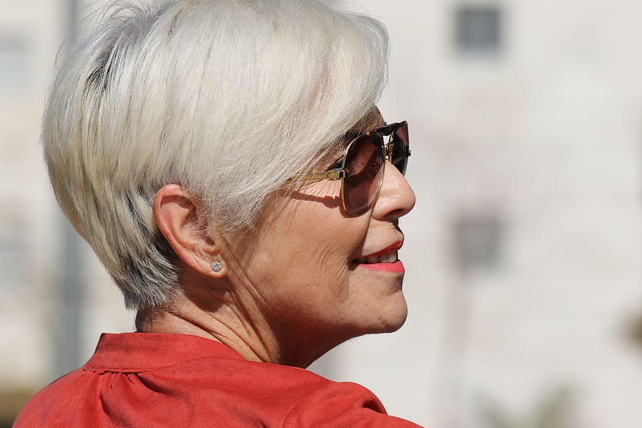 Makeup Tips for Gray Hair: Image shows a middle aged woman with a short gray hairstyle wearing sunglasses, coral lip color and an orange blouse. 