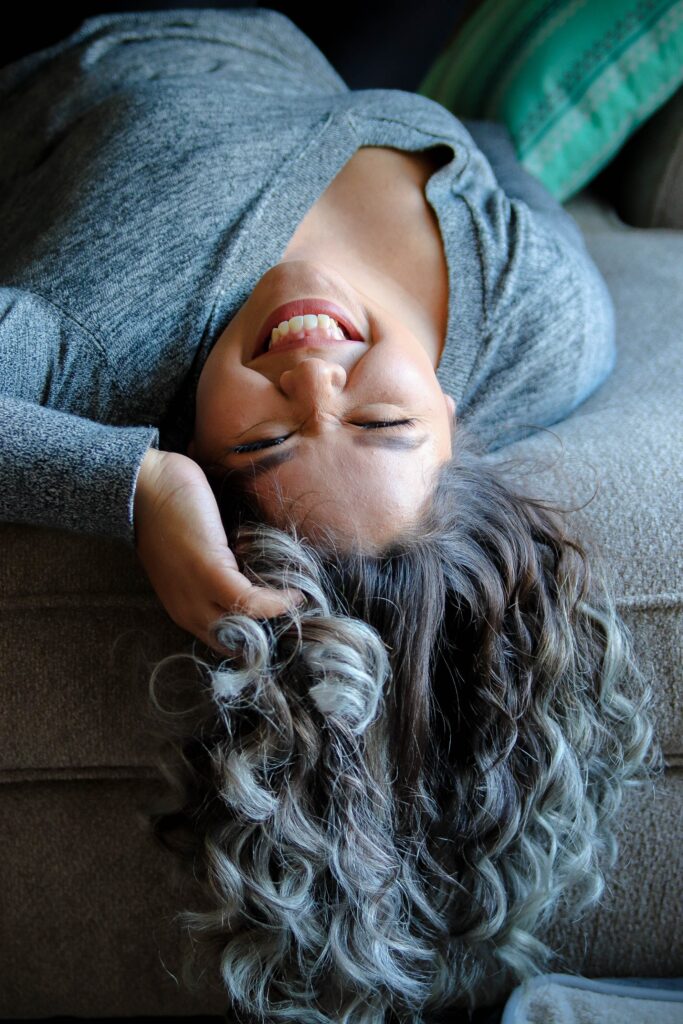 Makeup tips for gray hair. Image is of a woman lying on her back, gray curly hair dangling off a couch. She has a happy expression on her face. 